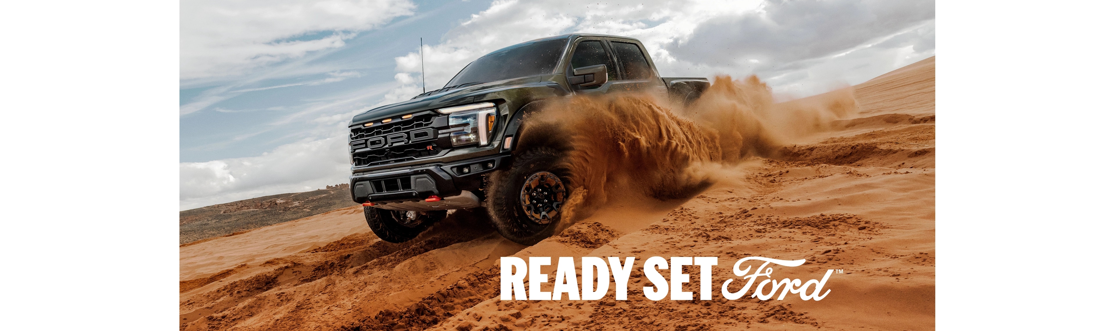 Three-quarter front view of a dark green Ford F-150 Raptor driving off-road through deep sand. White text “Ready Set Ford” appears in the lower left-hand corner. 