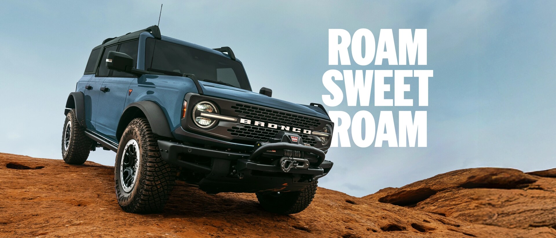 A three-quart front view of a 2025 blue-grey Ford Bronco descending rugged terrain. White “Roam Sweet Roam” text appears beside the Bronco.
