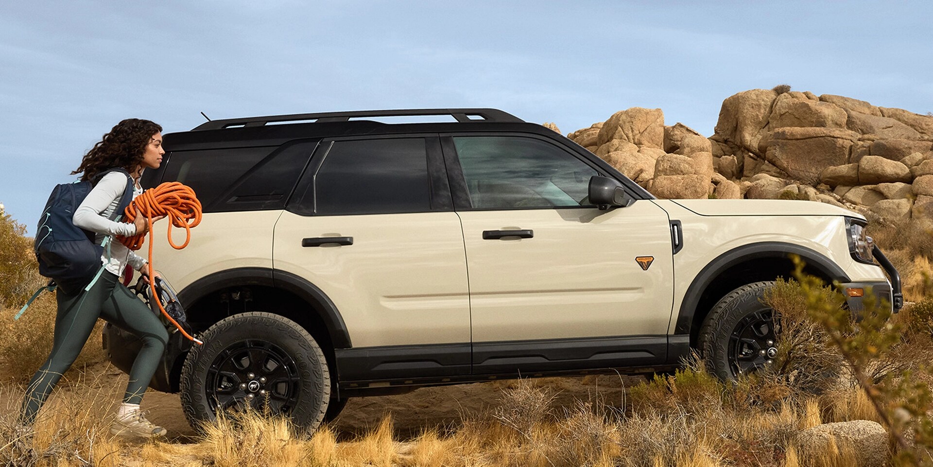 A female climber approaching a white Bronco Sport with boulders in the background. 