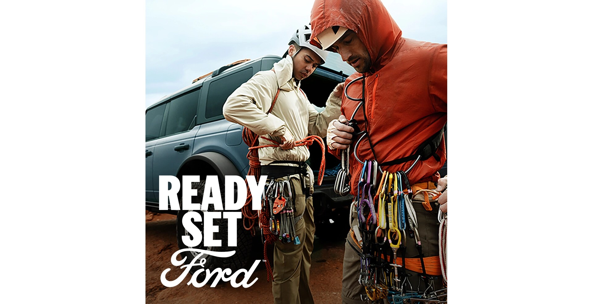 Two male rock climbers prepare for a climb beside a grey-blue Ford Bronco. White text “Ready Set Ford” is in the lower left corner.