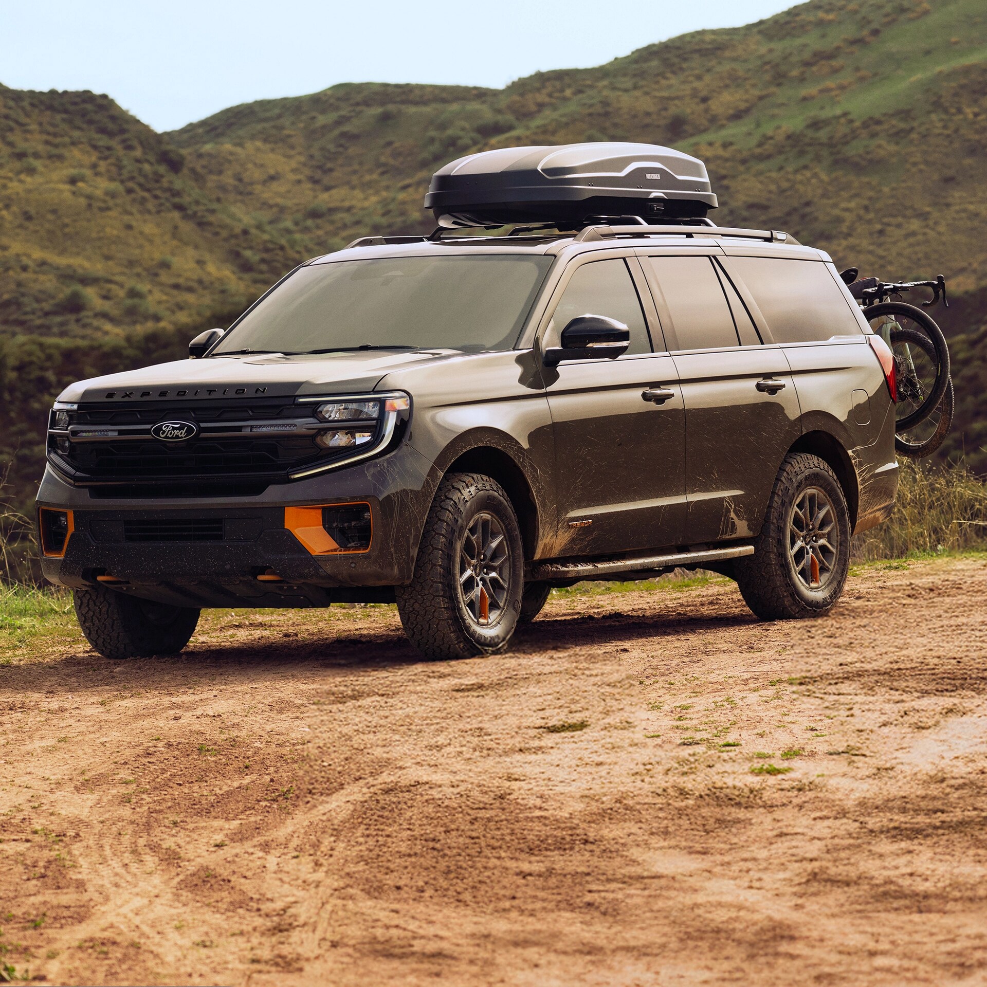 Three-quarter front view of a dark green 2025 Ford Expedition Tremor, with cargo accessory on roof, parked on a dirt trail in a hilly terrain. 
