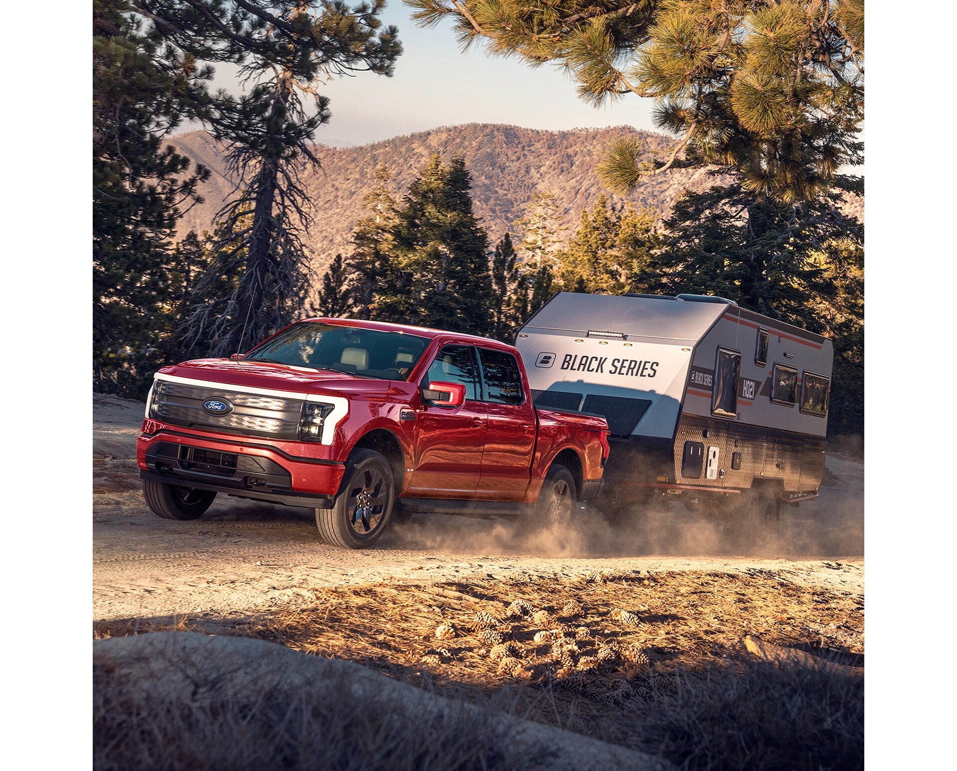 A three-quarter profile view of a red F-150 Lightning towing an RV trailer through a forested trail in a mountainous setting. 