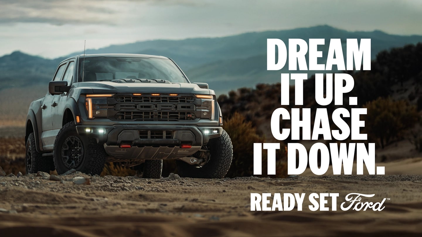 Three-quarter front view of a Ford F-150 Raptor parked on dirt with mountains in the background. "Dream it up. Chase it down. Ready Set Ford."