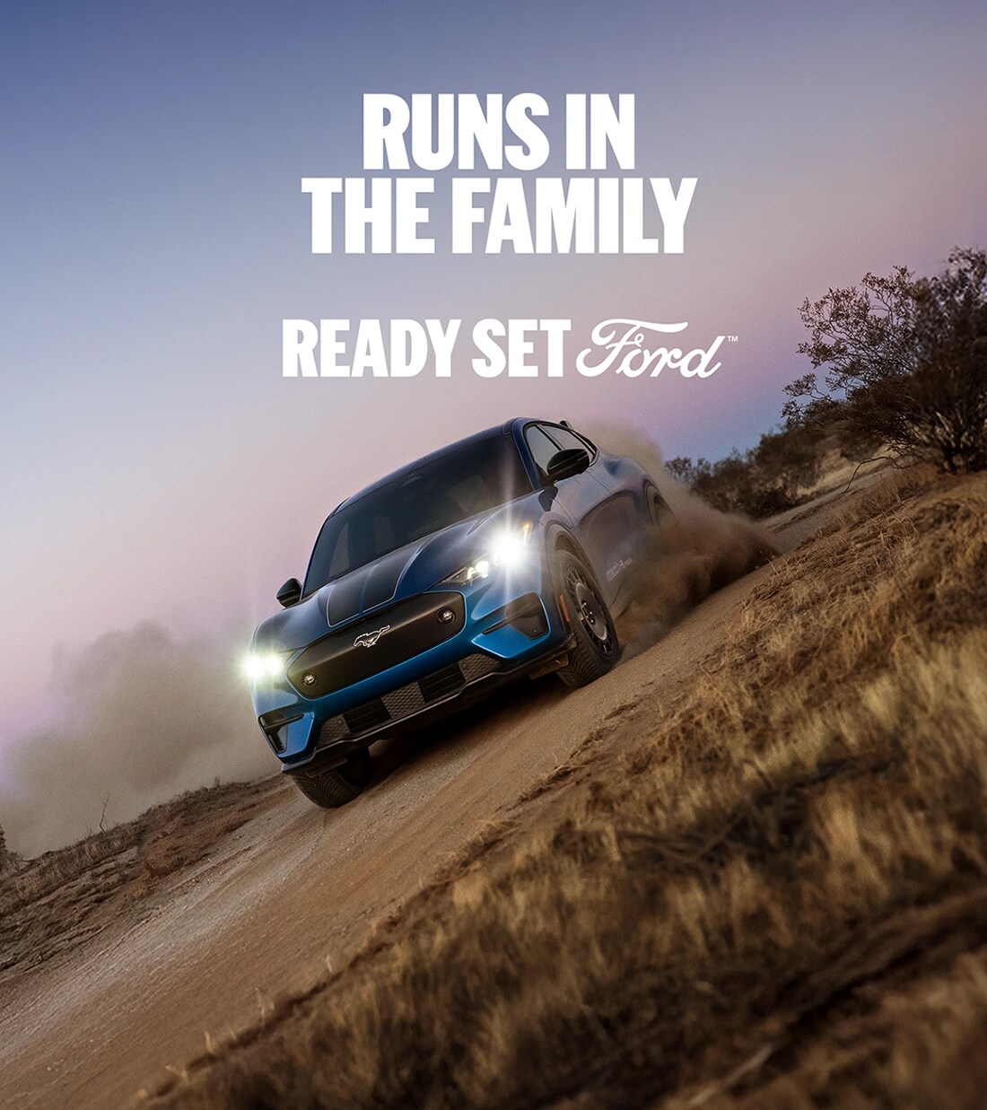 Three-quarter front view of a blue Ford Mustang Mach-E electric SUV driving on a dirt road at an angle. "Runs in the family. Ready Set Ford."