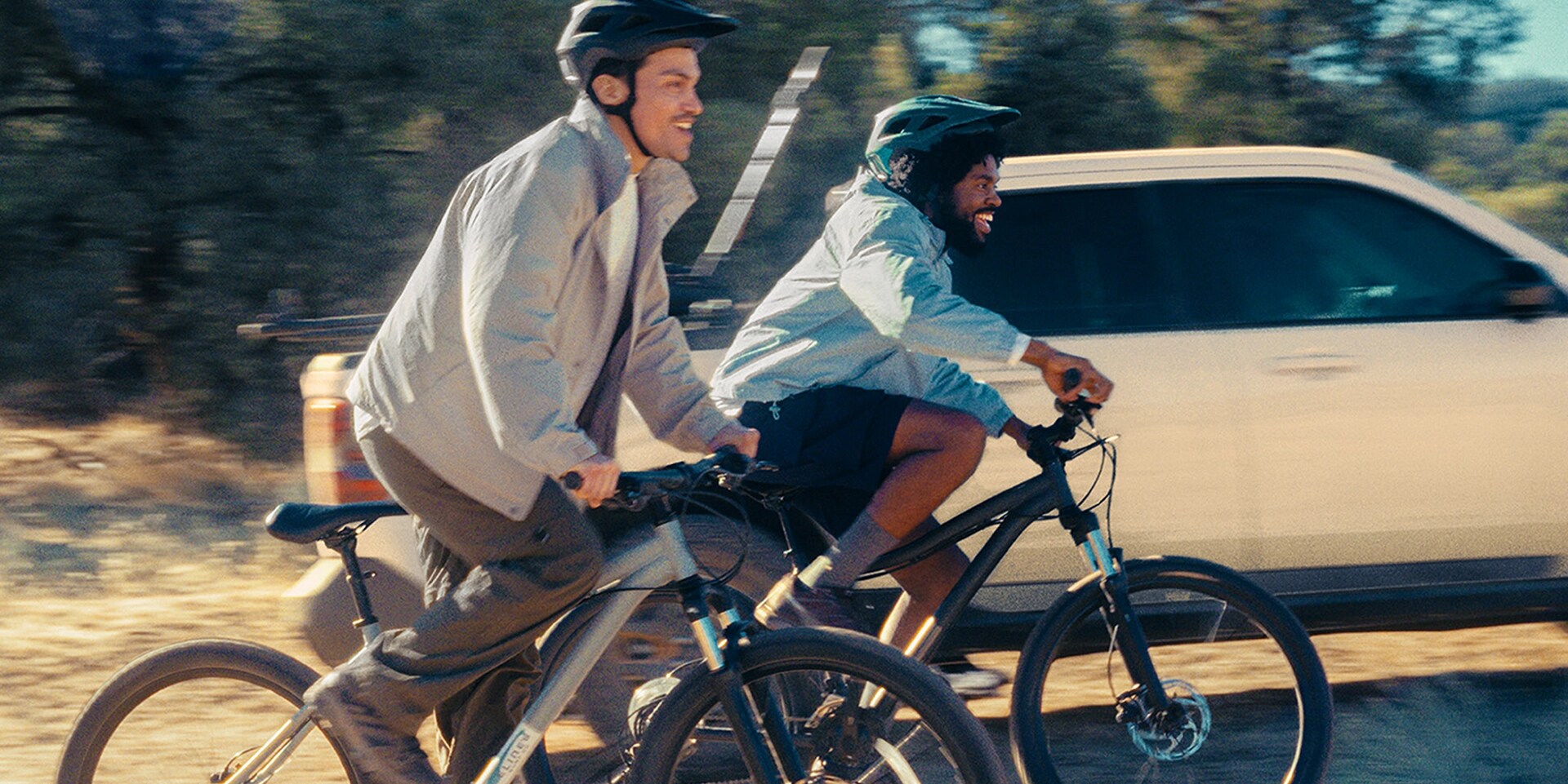 Two cyclists next to a white Ford Maverick on a dirt road. 