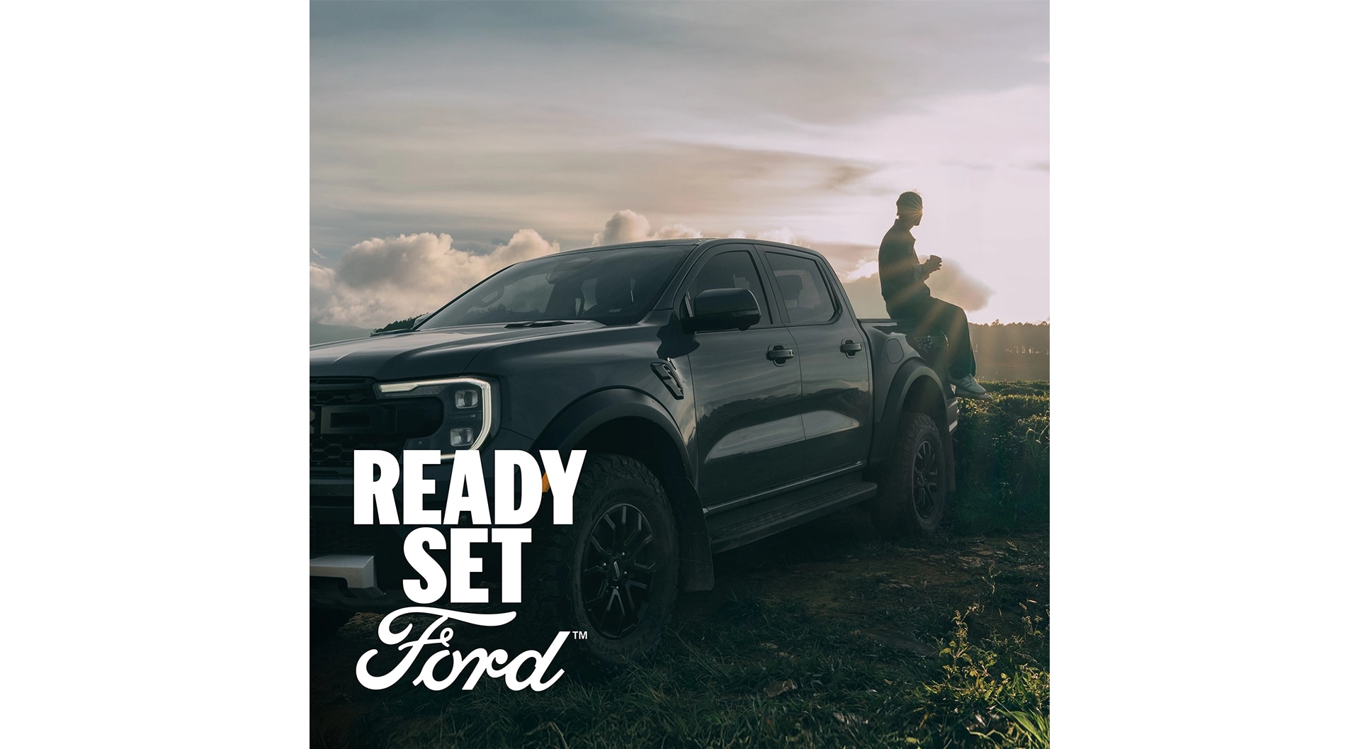 Three-quarter view of a grey Ford Ranger Raptor parked on grass. A person sits on the end of the truck bed, the sun shining in the background. White text “Ready Set Ford” appears in the lower-left corner.
