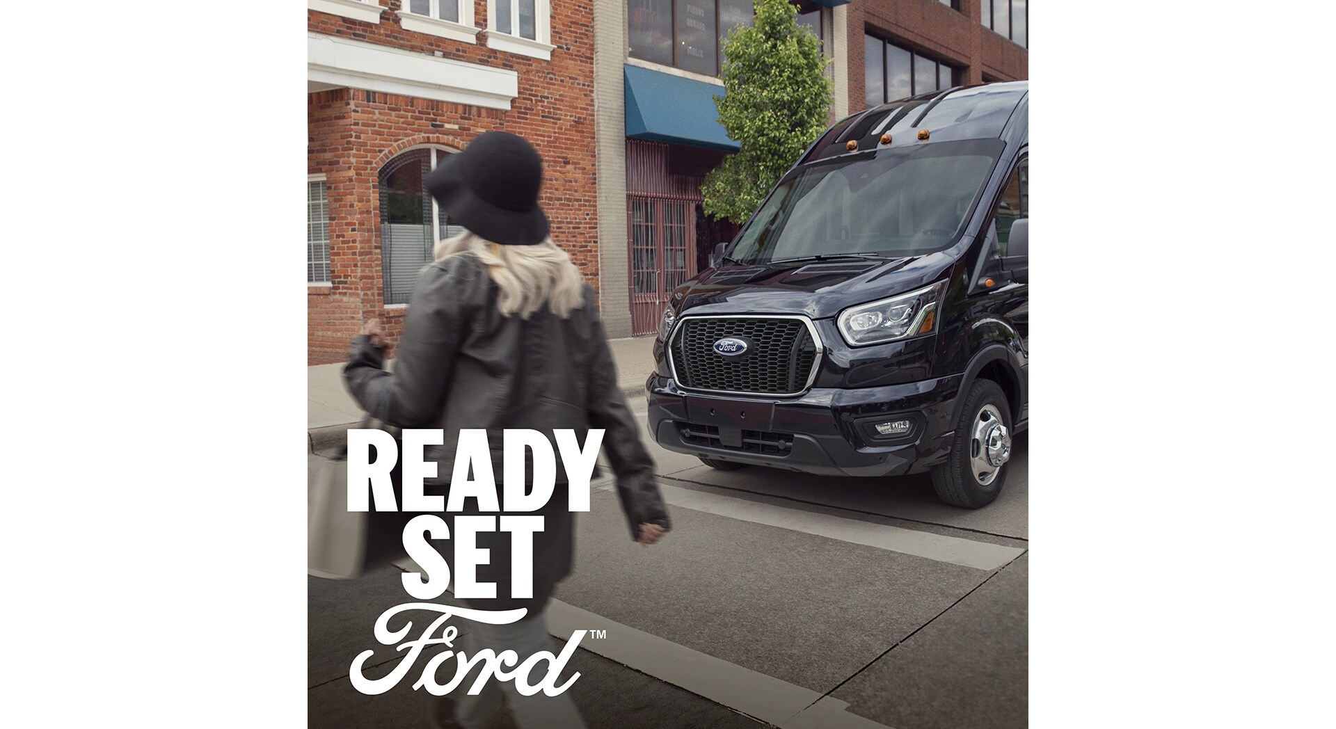 A three-quarter front view of a 2025 Ford Transit driving in an urban setting. In the lower left hand corner white text appears, “Ready Set Ford”. 