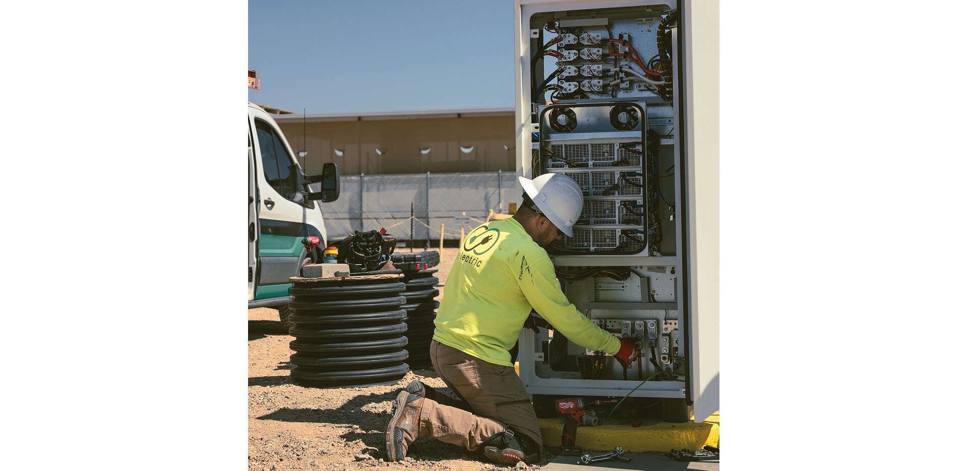 A male construction worker works on an electrical system at a construction site, with a Ford Transit partially visible on the right. 