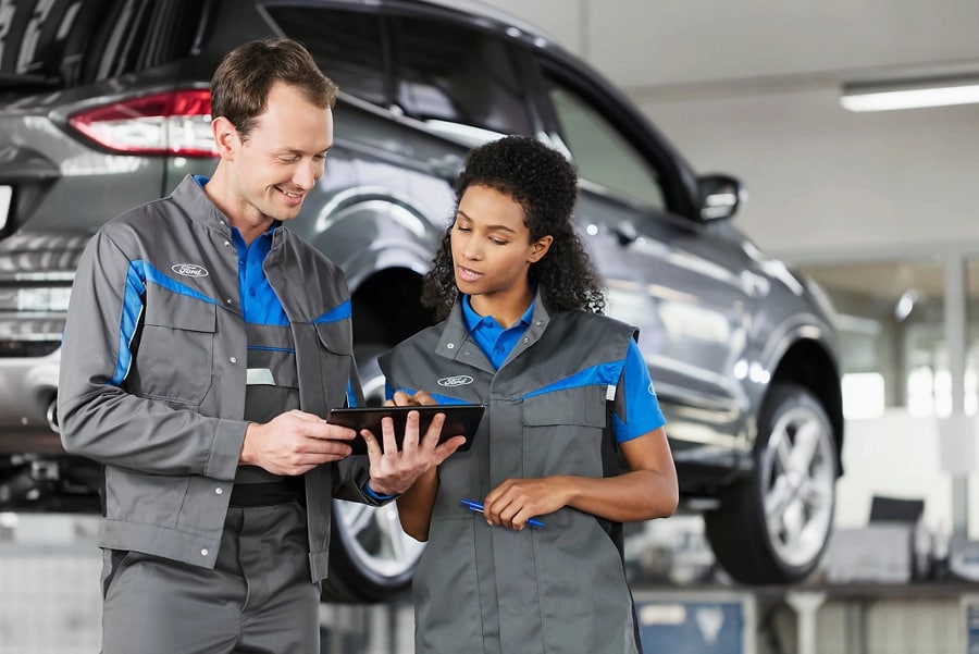 Two Ford employees in front of a vehicle on a car lift looking at a tablet.
