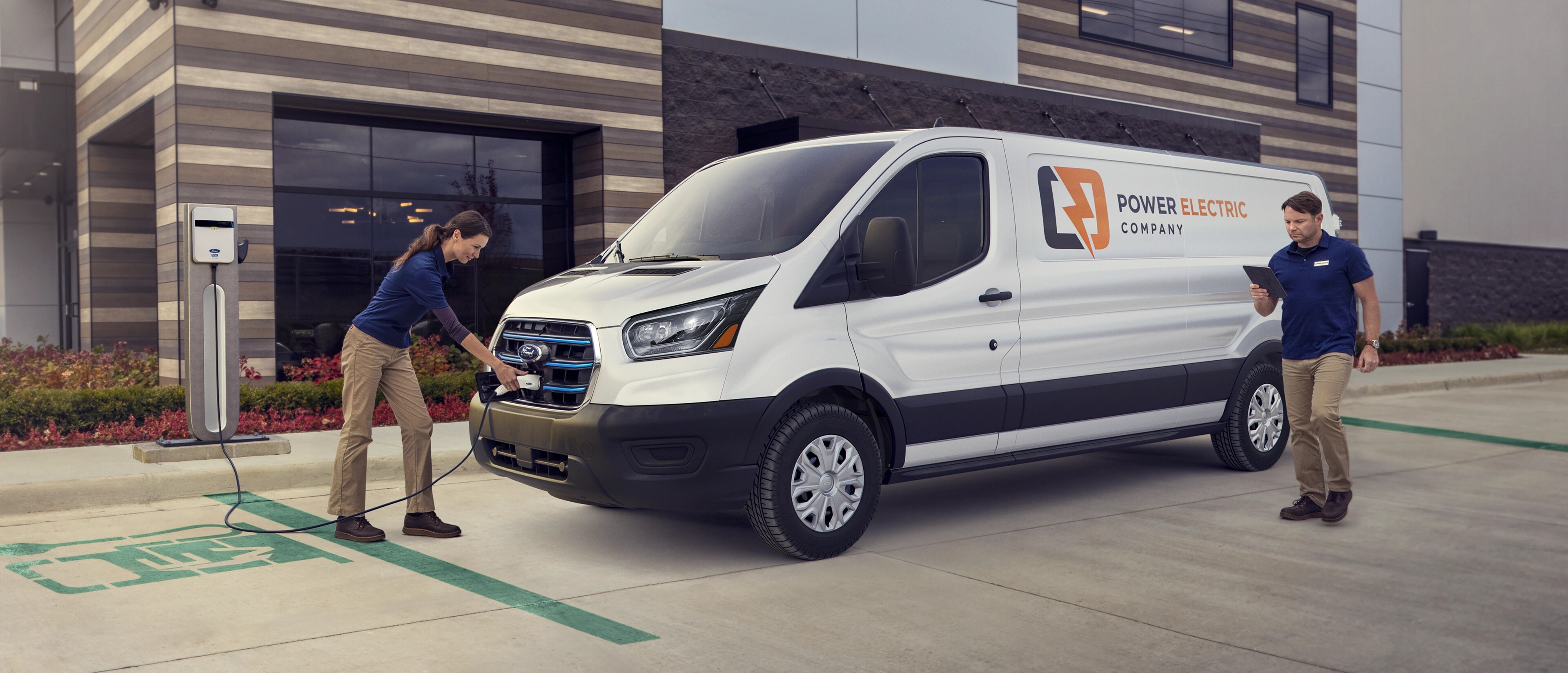 A white 2025 Ford E-Transit™ van parked and being charged by two workers outside a business