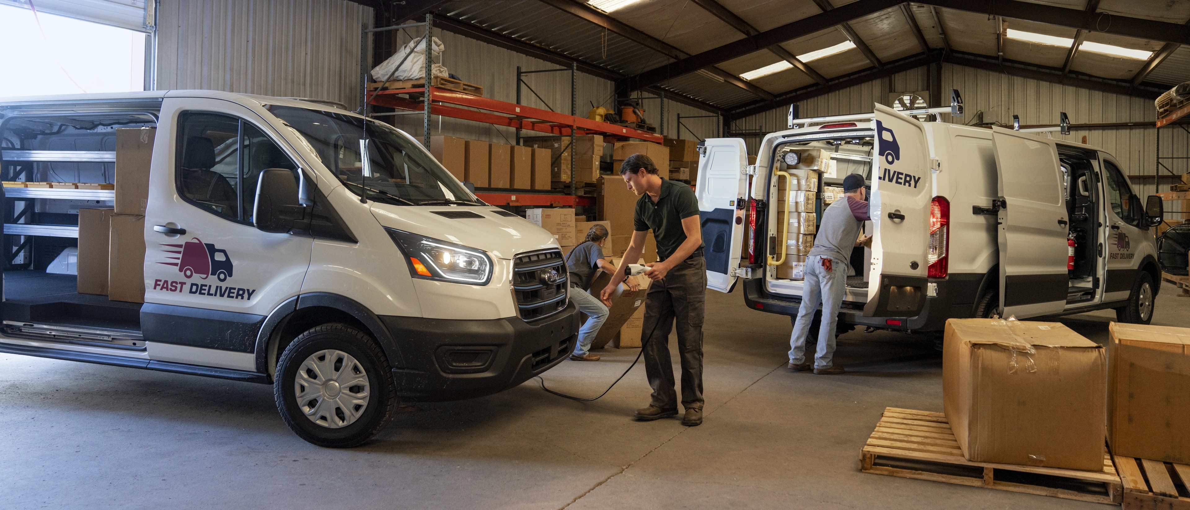 A white 2025 Ford E-Transit™ van parked with side door open while workers in a warehouse load cargo
