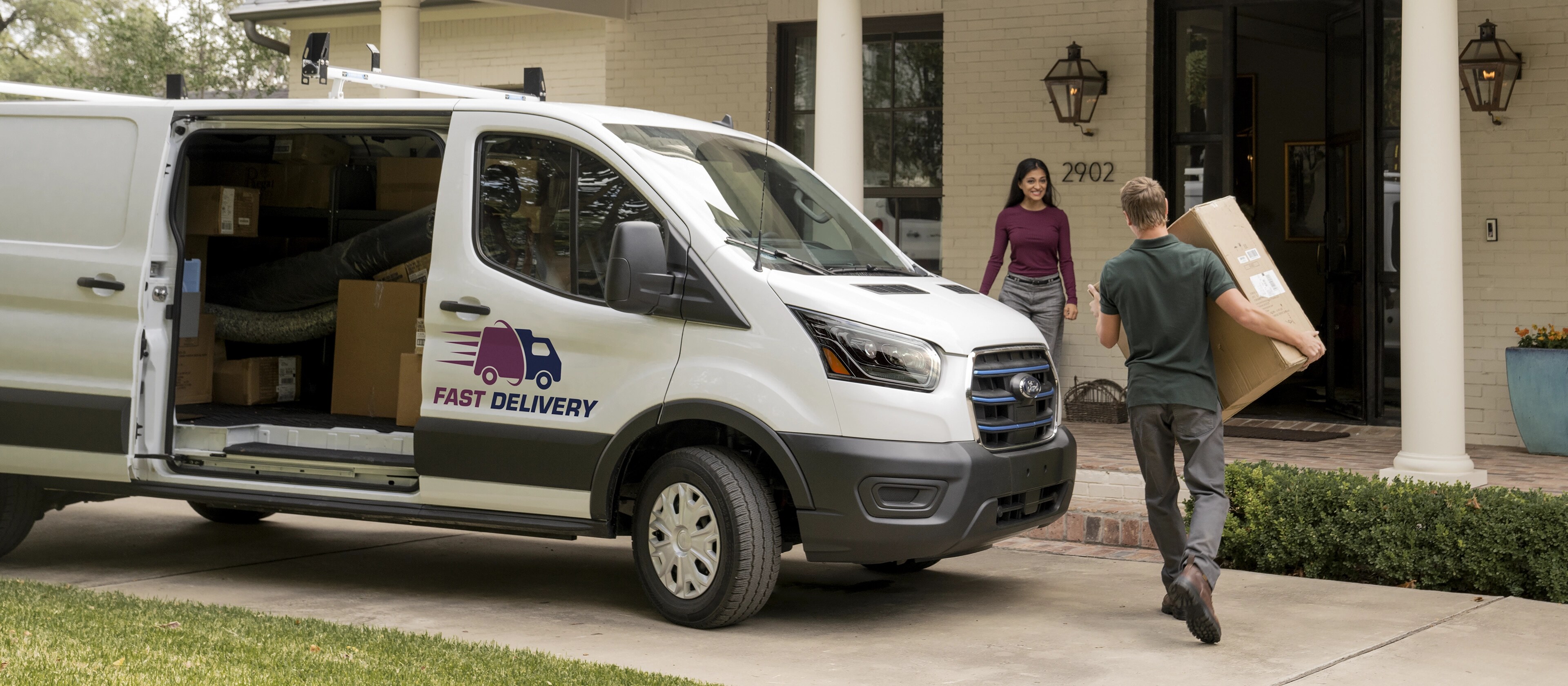 A white 2025 Ford E-Transit™ van parked as its driver walks to a house for delivery