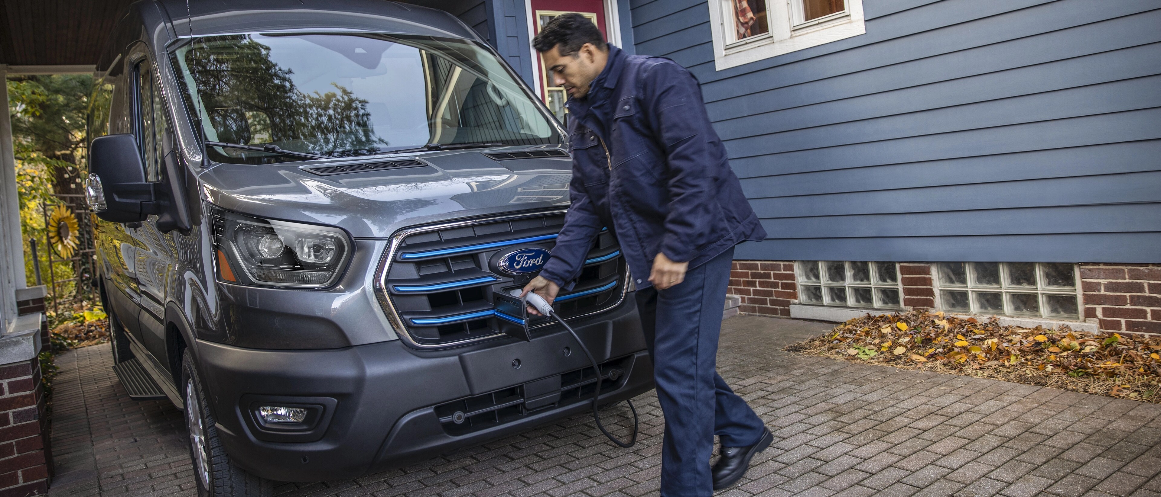 A grey 2025 Ford E-Transit™ van parked in a home driveway being charged for the night