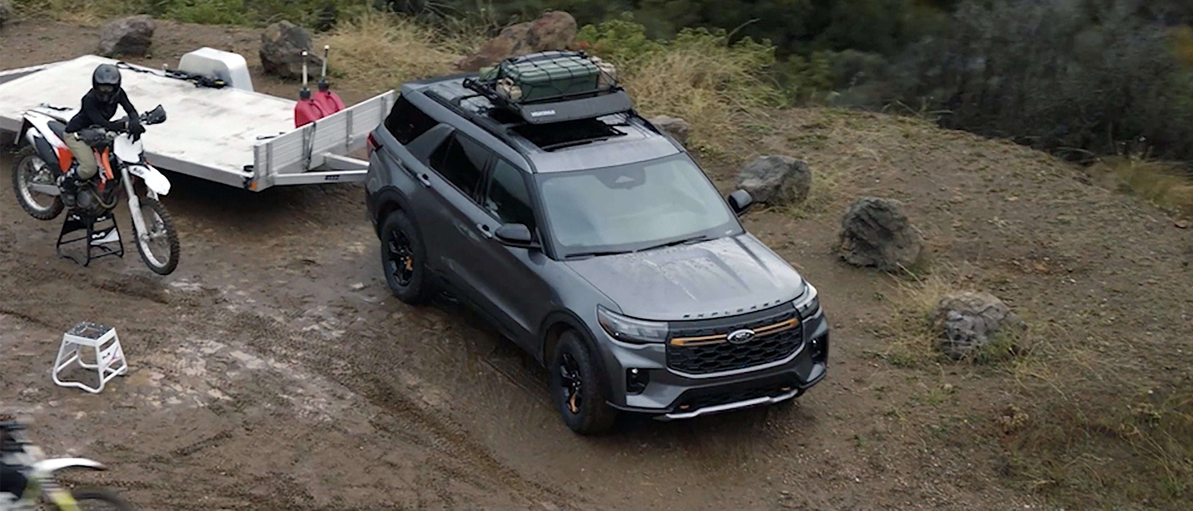 A 2026 Ford Explorer® Tremor® SUV with a trailer parked at a mountain overlook with a person riding a dirtbike nearby