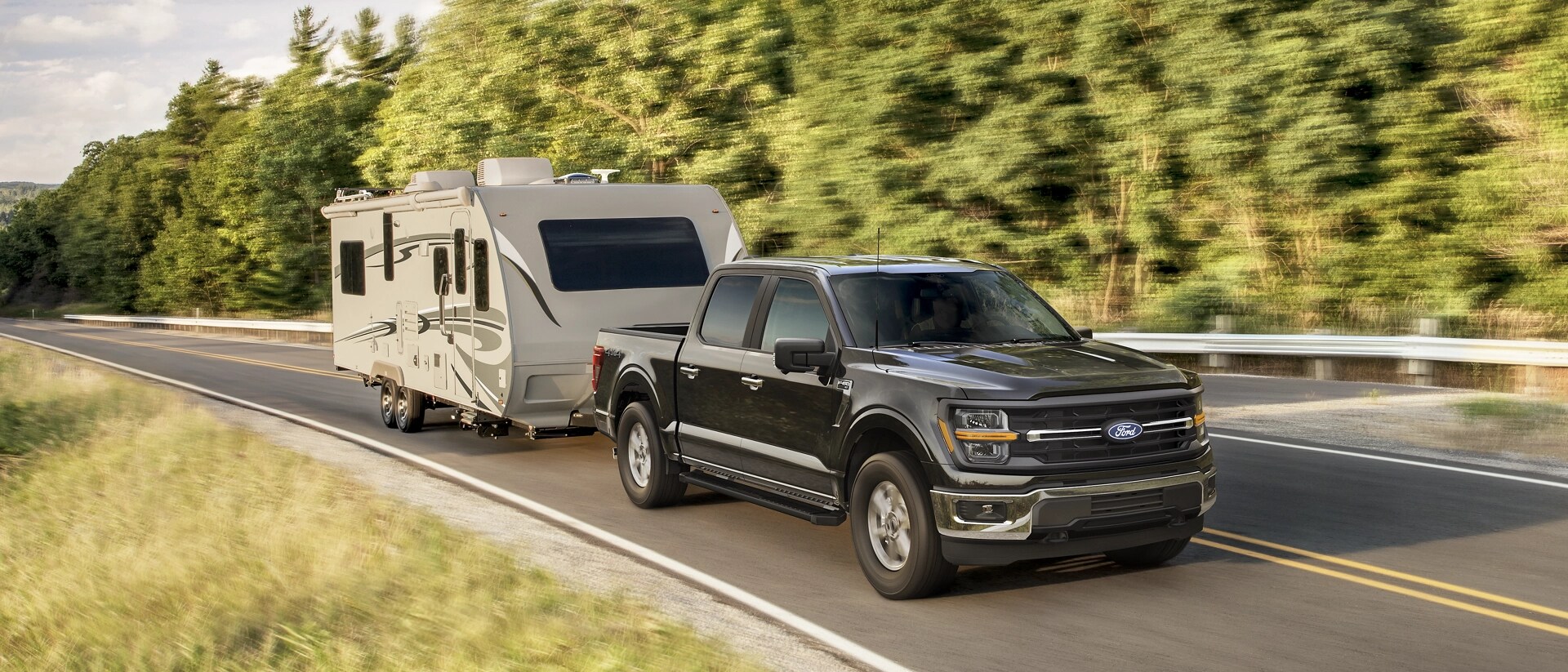 2025 Ford F-150® XLT model in Darkened Bronze Metallic pulling a camper on a tree-lined road