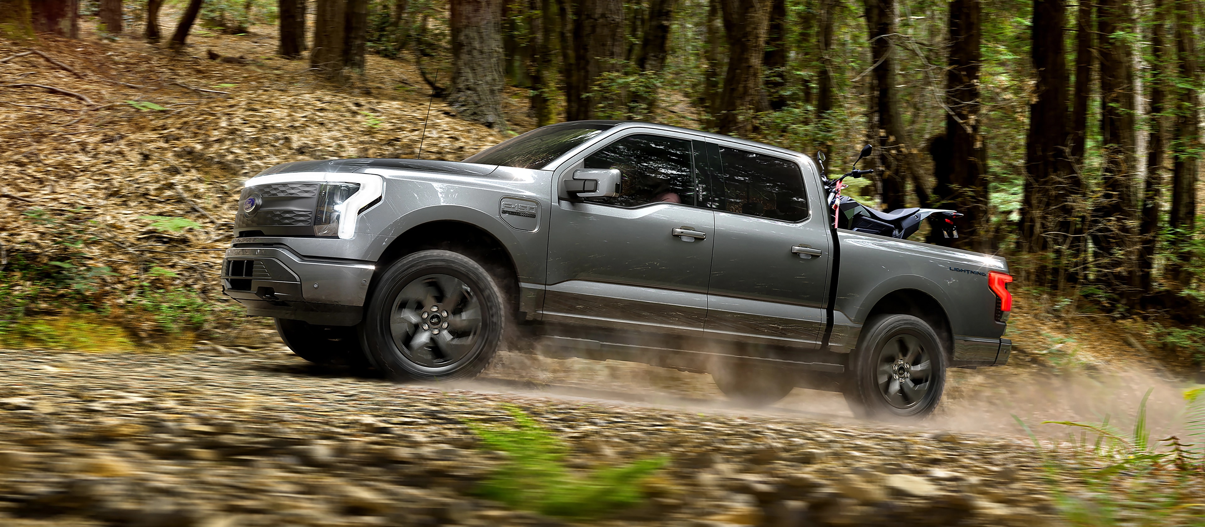 A 2025 Ford F-150® Lightning® pickup driving on a dusty road through the forest with an ATV secured in the cargo bed