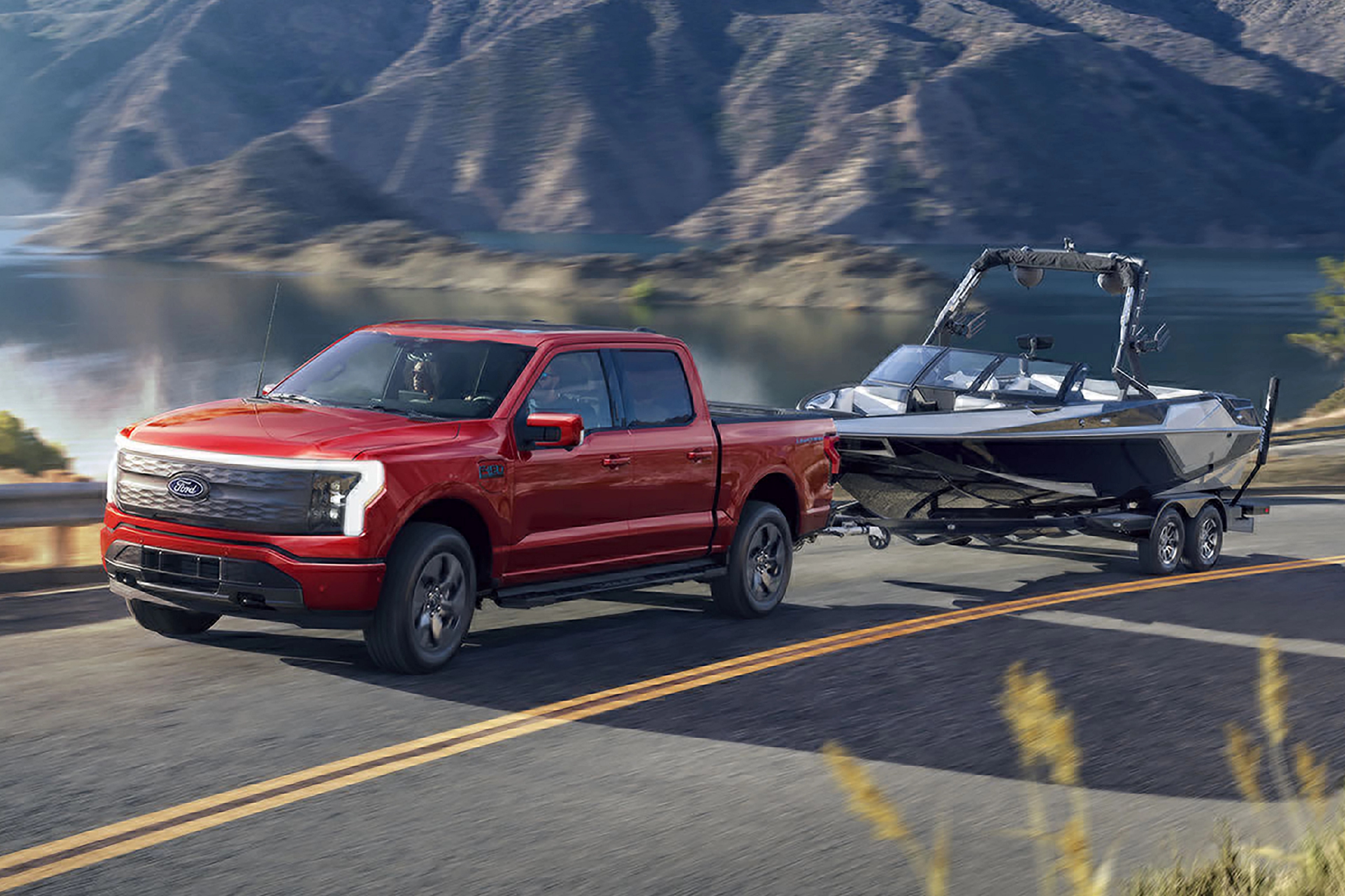 A 2025 Ford F-150® Lightning® hauling a boat on a trailer on a highway near a mountain lake