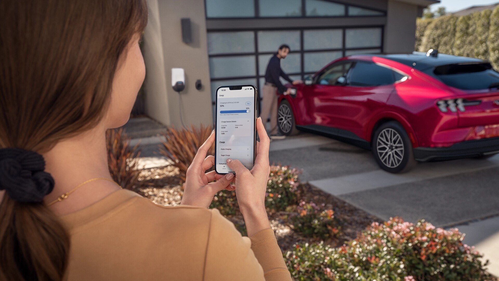 Woman using a phone with person plugging in a 2025 Ford Mustang Mach-E® in a driveway