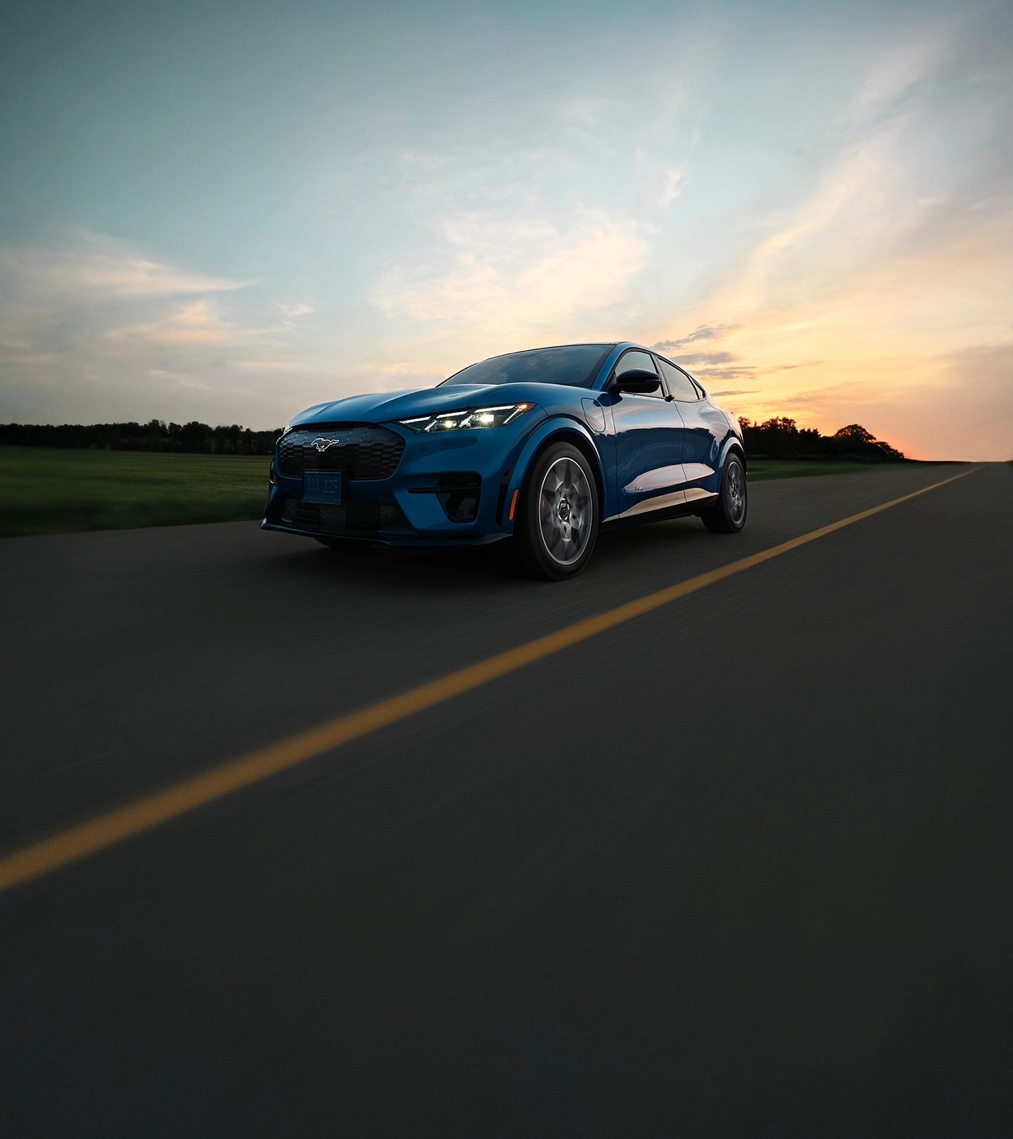 A blue 2025 Ford Mustang Mach-E® GT driving on an open road at sunset, with dramatic clouds and fading sunlight in the background.