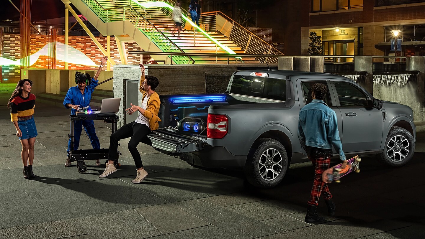 Young people relaxing near a 2025 Ford Maverick® pickup as a DJ plays music through speakers plugged in to a cargo bed outlet