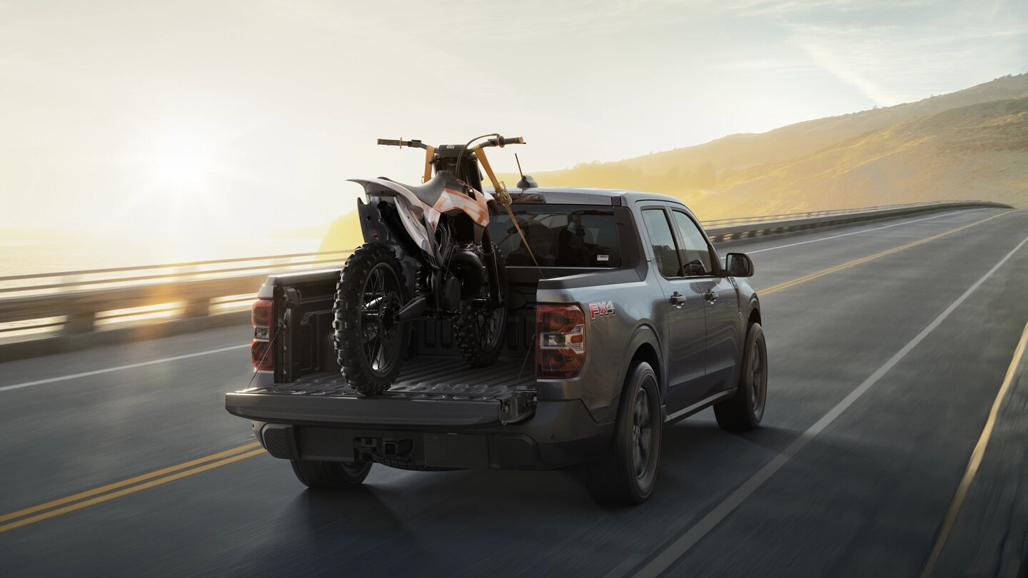 A 2025 Ford Maverick® pickup with a dirtbike secured in the bed, driving on a country highway at sunrise