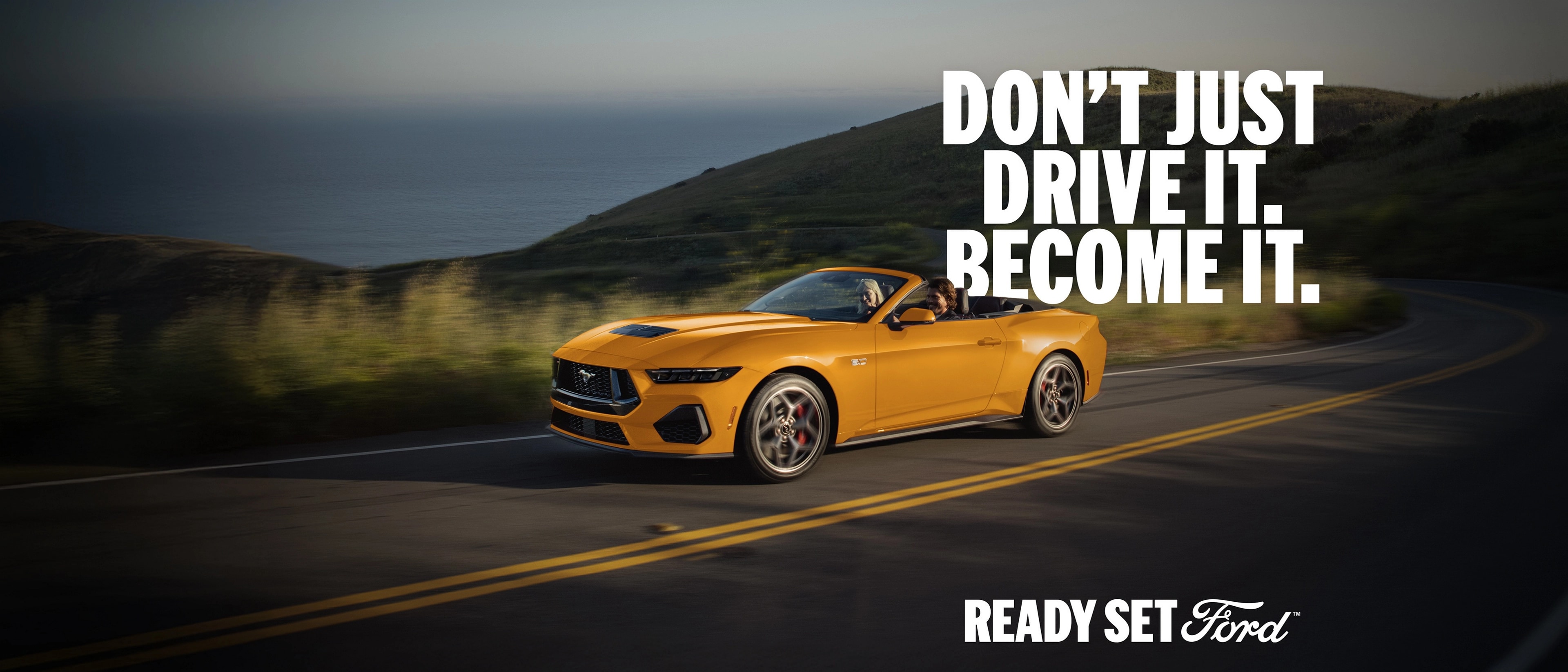 Two people in a 2026 Ford Mustang® GT Convertible in Orange Fury Metallic Tri-coat being driven on a coastal highway