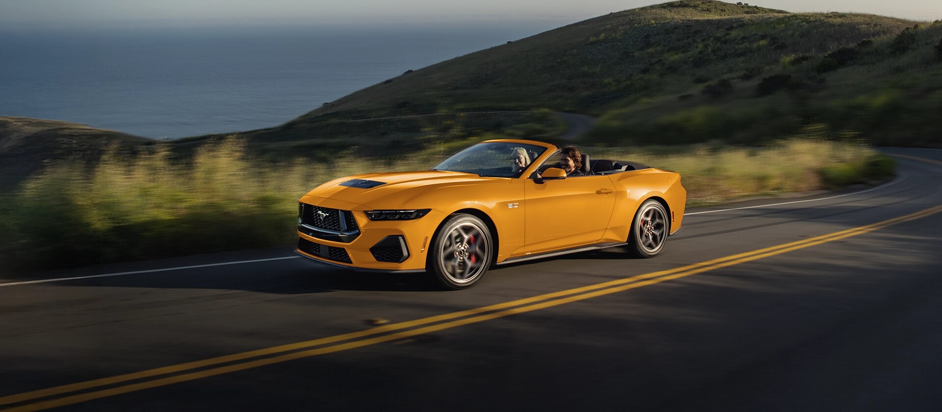 Two people in a 2026 Ford Mustang® GT convertible in Orange Fury Metallic Tri-coat being driven on a coastal highway