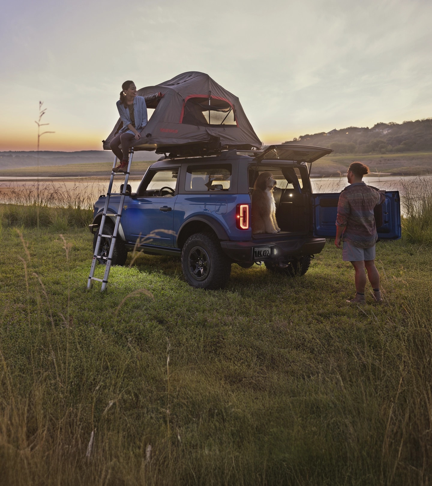 Two people and a dog around a Bronco with a roof-mounted tent.