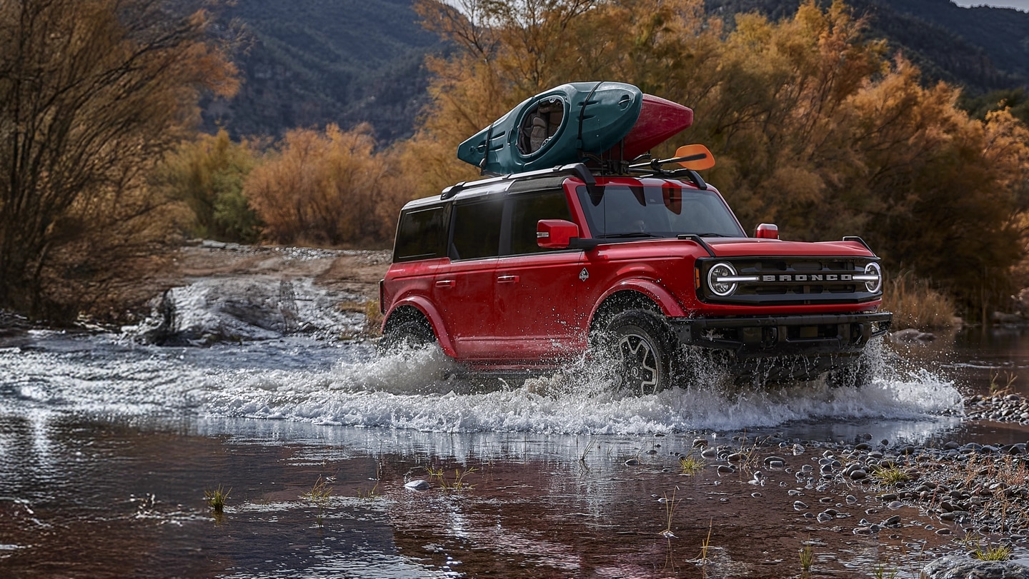 Bronco with roof-mounted canoe driving through water.