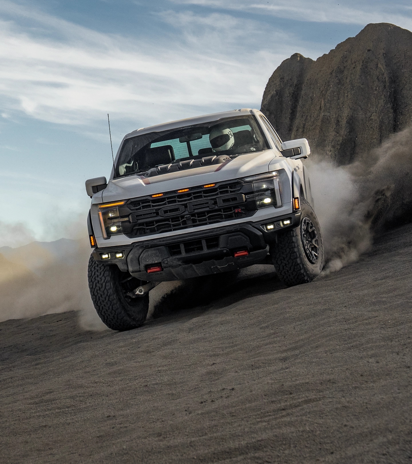 Ford F-150® Raptor® driving off-road, kicking up dust on a rocky hill under a blue sky.