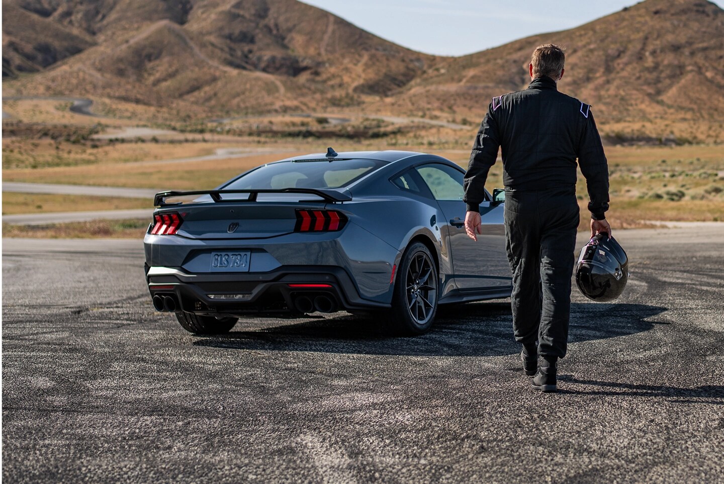 Person holding a racing helmet walks toward a Mustang Dark Horse parked on a track.