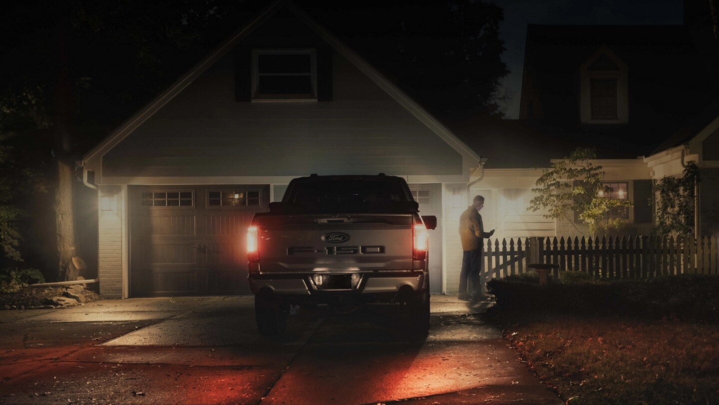 Person holding a smartphone standing next to an F-150 Lightning in a residential driveway.