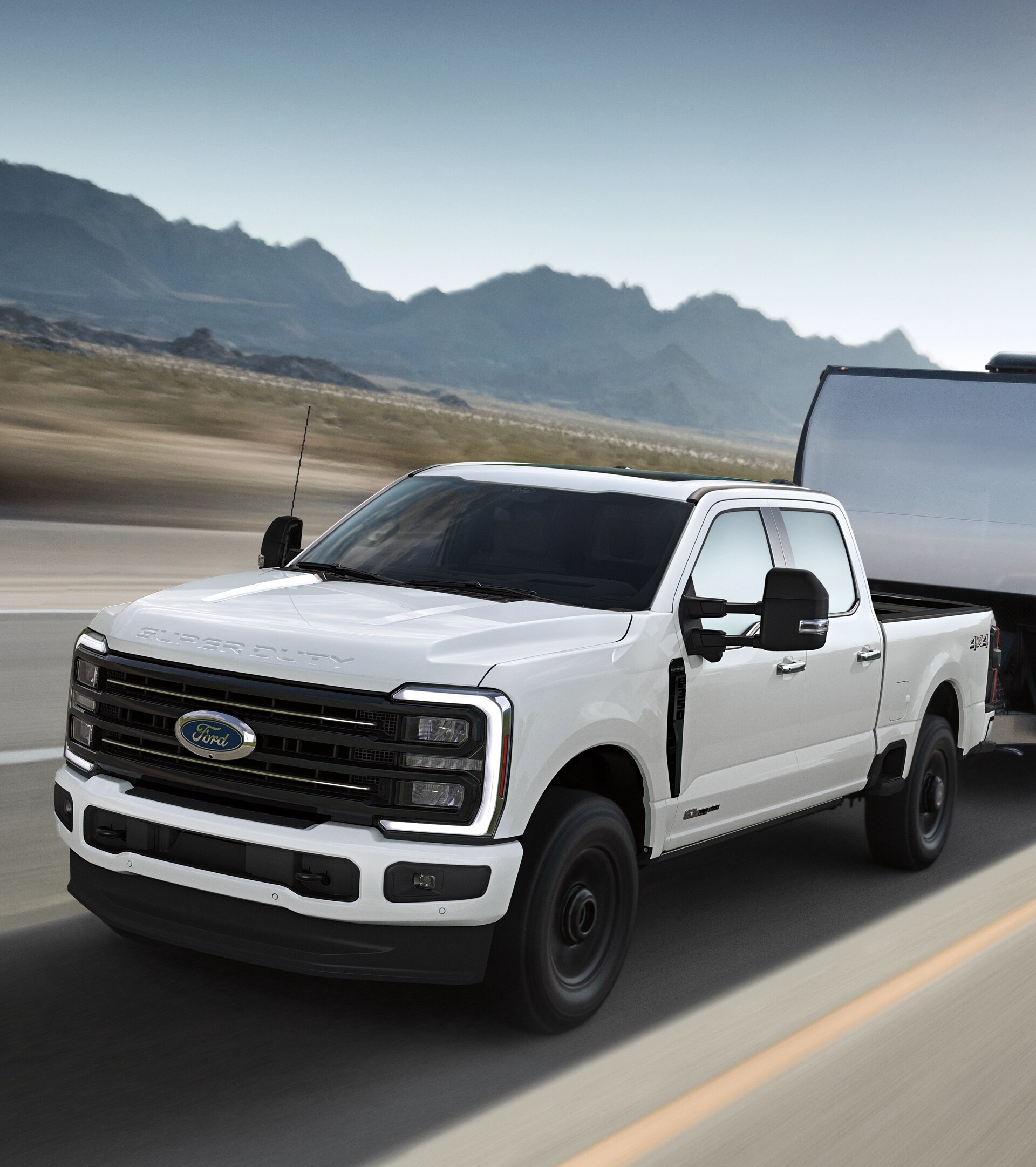 2026 Ford Super Duty® F-350® Platinum® model in Star White driving on a paved road while towing a large silver trailer