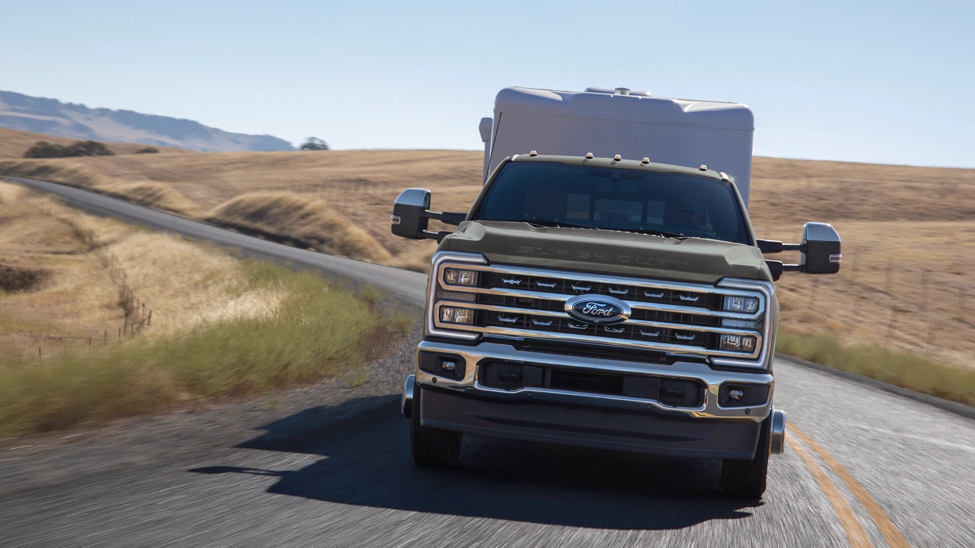 2026 Ford Super Duty® F-350® Lariat® model with the Chrome Package driving on a hilly paved road while towing a large trailer