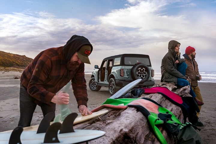 4-door 2023 Ford Bronco® Outer Banks™ model in Cactus Grey parked on the beach with 3 surfers