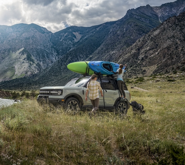 A person is standing outside a 2024 Ford Bronco® Sport SUV that is parked by a lake with a kayak on the roof