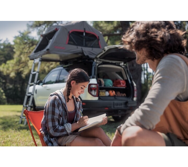 A young woman writes in her journal while a man sits near her with a 2024 Ford Bronco® Sport SUV in the background