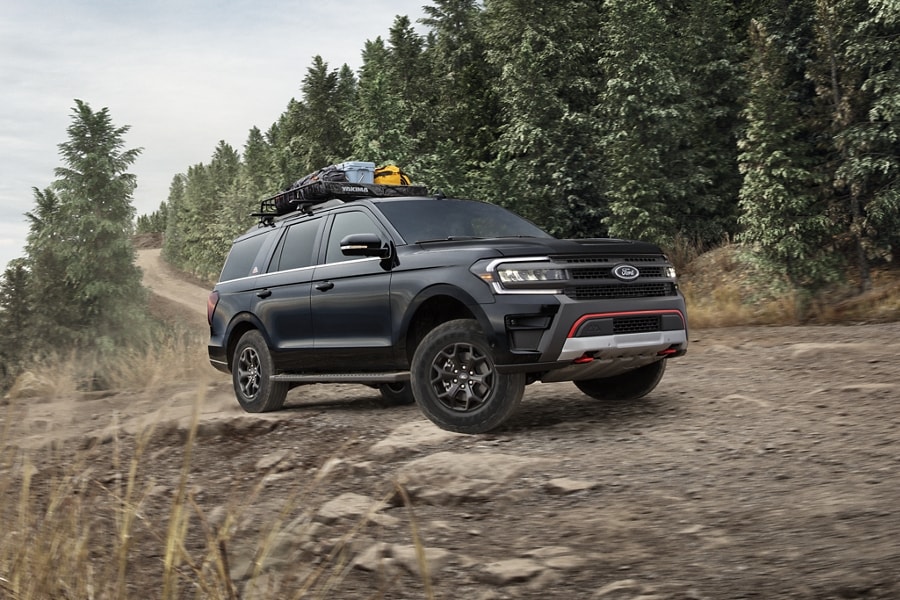 A 2023 Ford Expedition Timberline SUV with outdoor gear secured to roof parked on a dirt trail road with forest in the background
