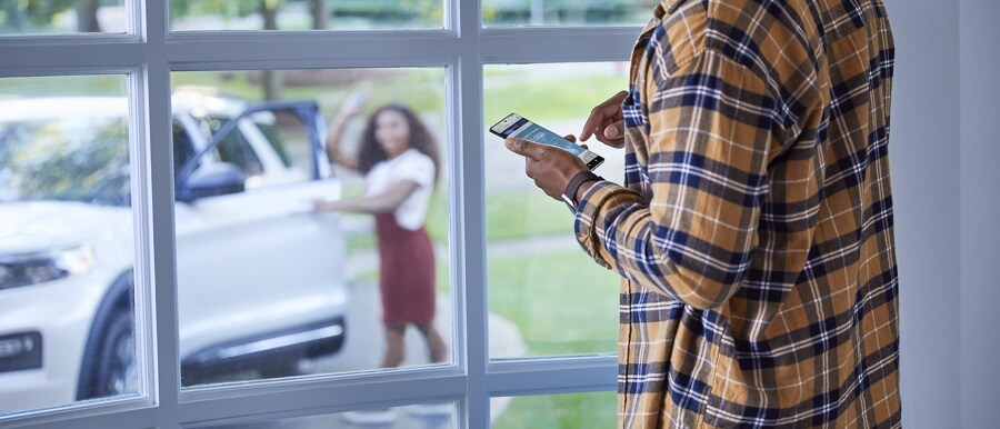 Person on a smartphone in a house using the FordPass™ App with a driver outside near a 2023 Ford Explorer® SUV