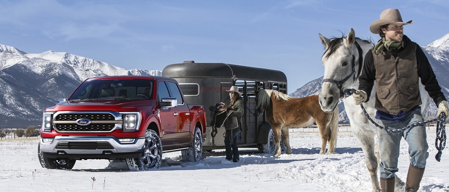 A man walking a horse near a 2023 Ford F-150® King Ranch™ in Rapid Red hitched to a horse trailer on a snow-covered plain