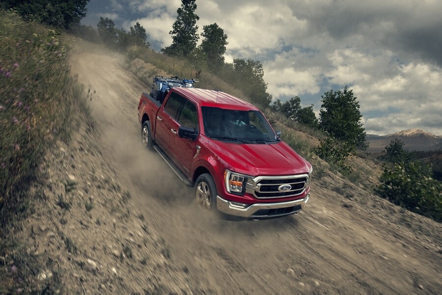 A 2023 Ford F-150® FX4 in Rapid Red Metallic Tinted Clearcoat being driven down a sloped dirt road kicking up dust