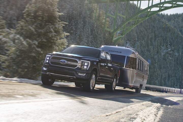 A 2023 Ford F-150® towing a trailer near a tree-lined mountain on a highway