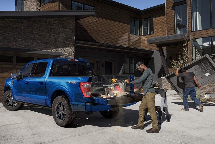 A man cutting wood with a power saw on the tailgate of a 2023 Ford F-150® LARIAT® in Atlas Blue in front of a modern home