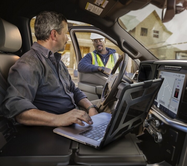 Man using the centre console of Interior Work Surface to work on a lap top while talking to a man standing outside the door