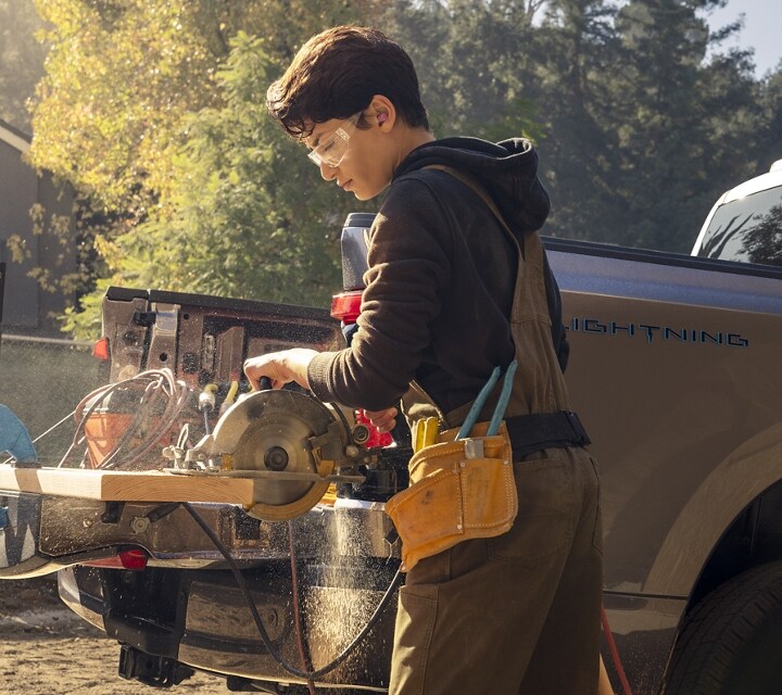 A construction worker using the 2023 Ford F-150® Lightning® flat work surface of the tailgate to cut a board