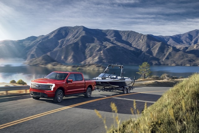 A 2023 Ford F-150® Lightning® towing a boat up a mountain road, lake and mountains in background