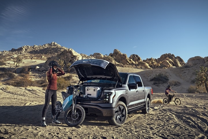 2023 Ford F-150® Lightning® open parked in rocky, desert terrain shown with frunk open and electric cyclists nearby