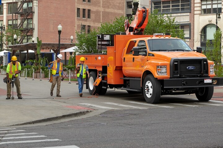 Workers walking near 2023 Ford F-750 Crew Cab with aerial lift upfit in Tangier Orange parked on city street