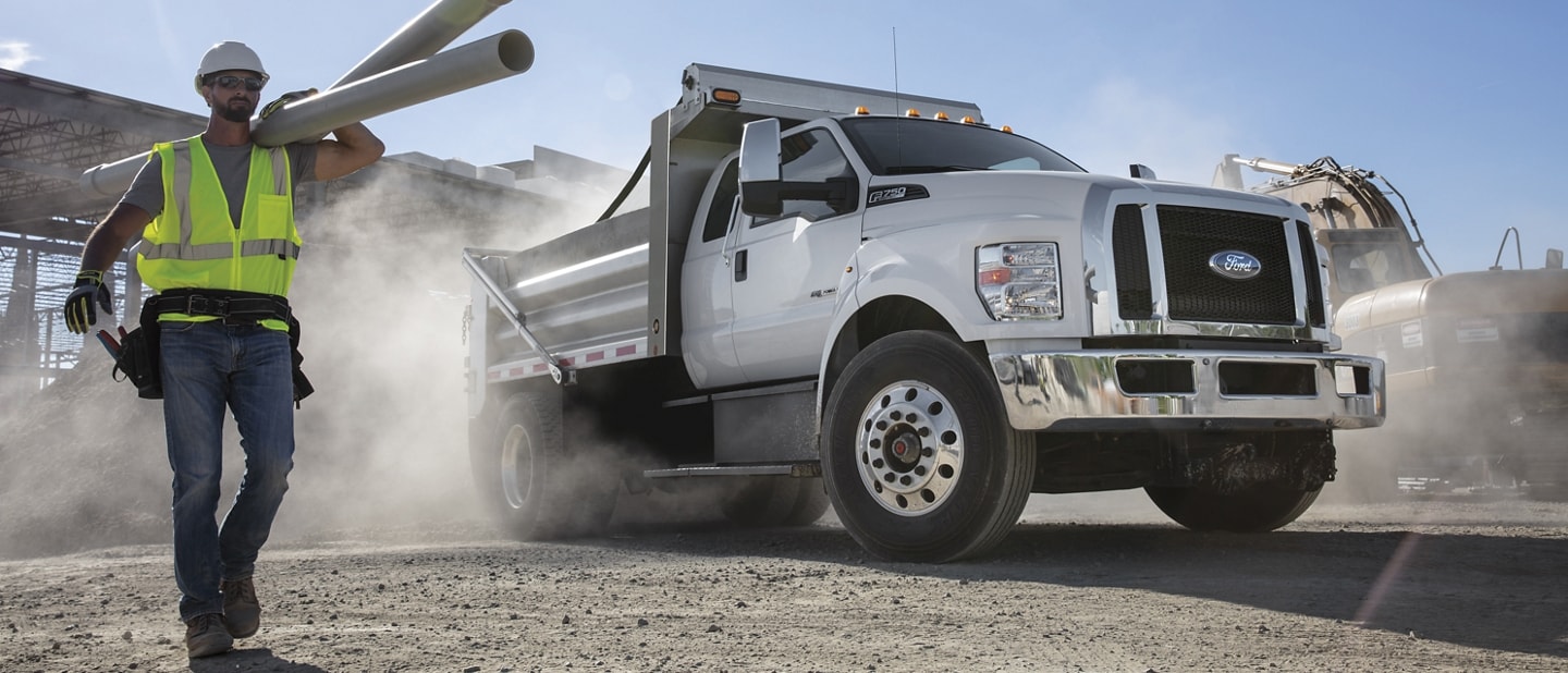 Worker carrying pipes on a dusty construction site next to a 2026 Ford F-750® with dump truck upfit shown in Oxford White