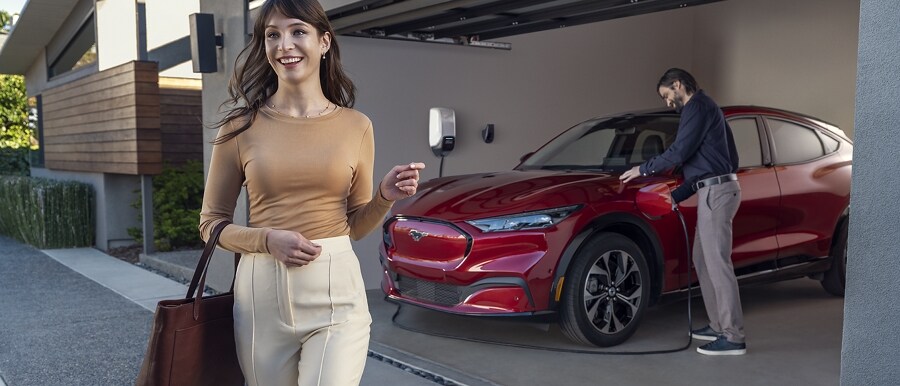 Woman walks in front of a 2023 Ford Mustang Mach-E® parked in a driveway with a man holding a Ford mobile charger